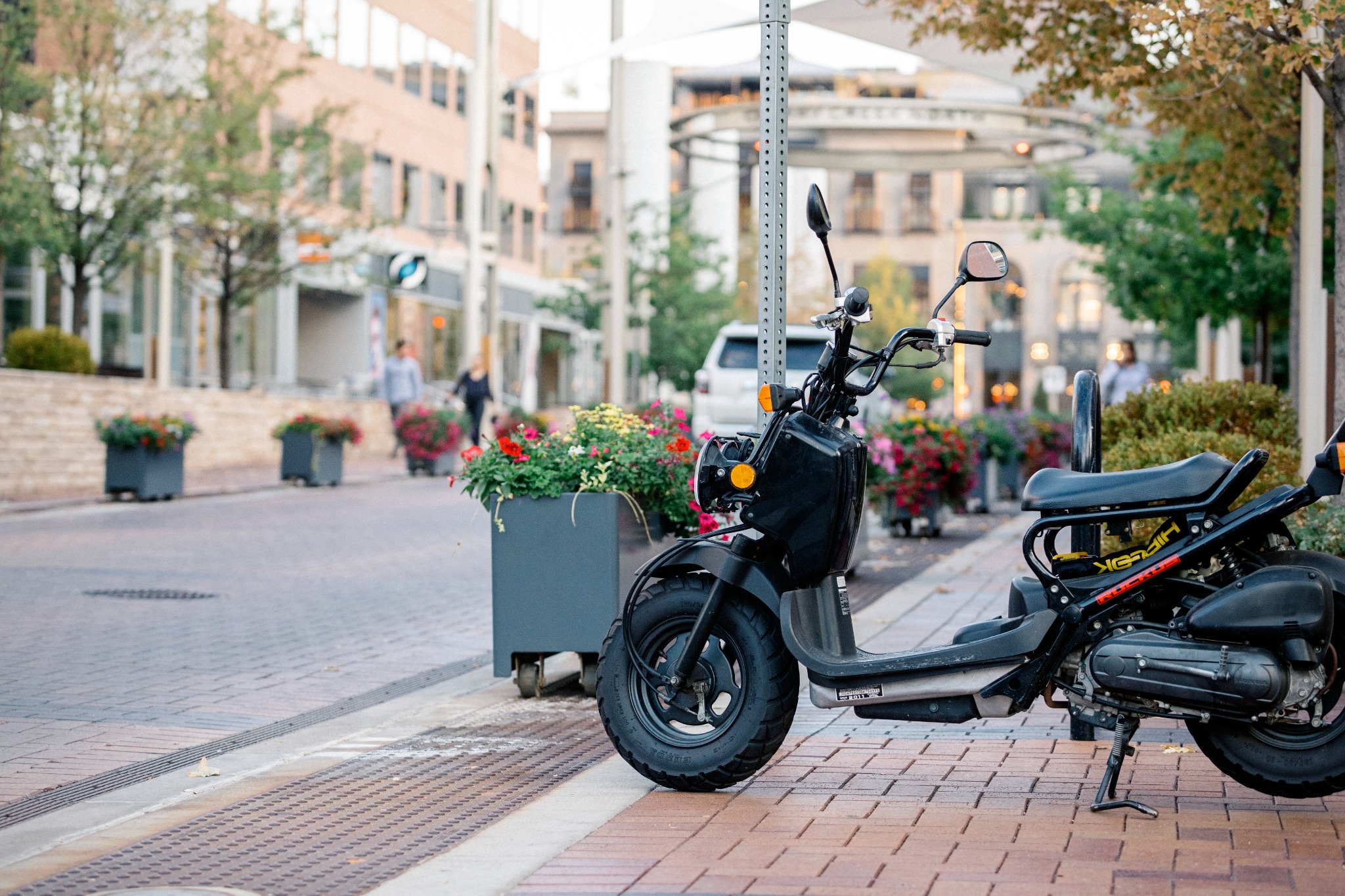A black motor scooter parked beside flower planters on a tree-lined shopping street.